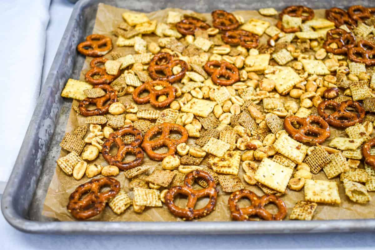 Pretzels, crackers, peanuts, and cereal spread on a parchment-lined sheet pan ready to bake.