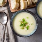 Bowl of creamy potato leek soup topped with green onions, served with sliced bread and a spoon on the side.