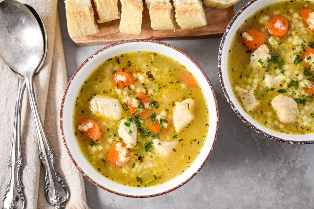 Overhead view of two bowls of the chicken soup with carrots and pasta, served with sliced bread, butter, and silver spoons on a neutral background.