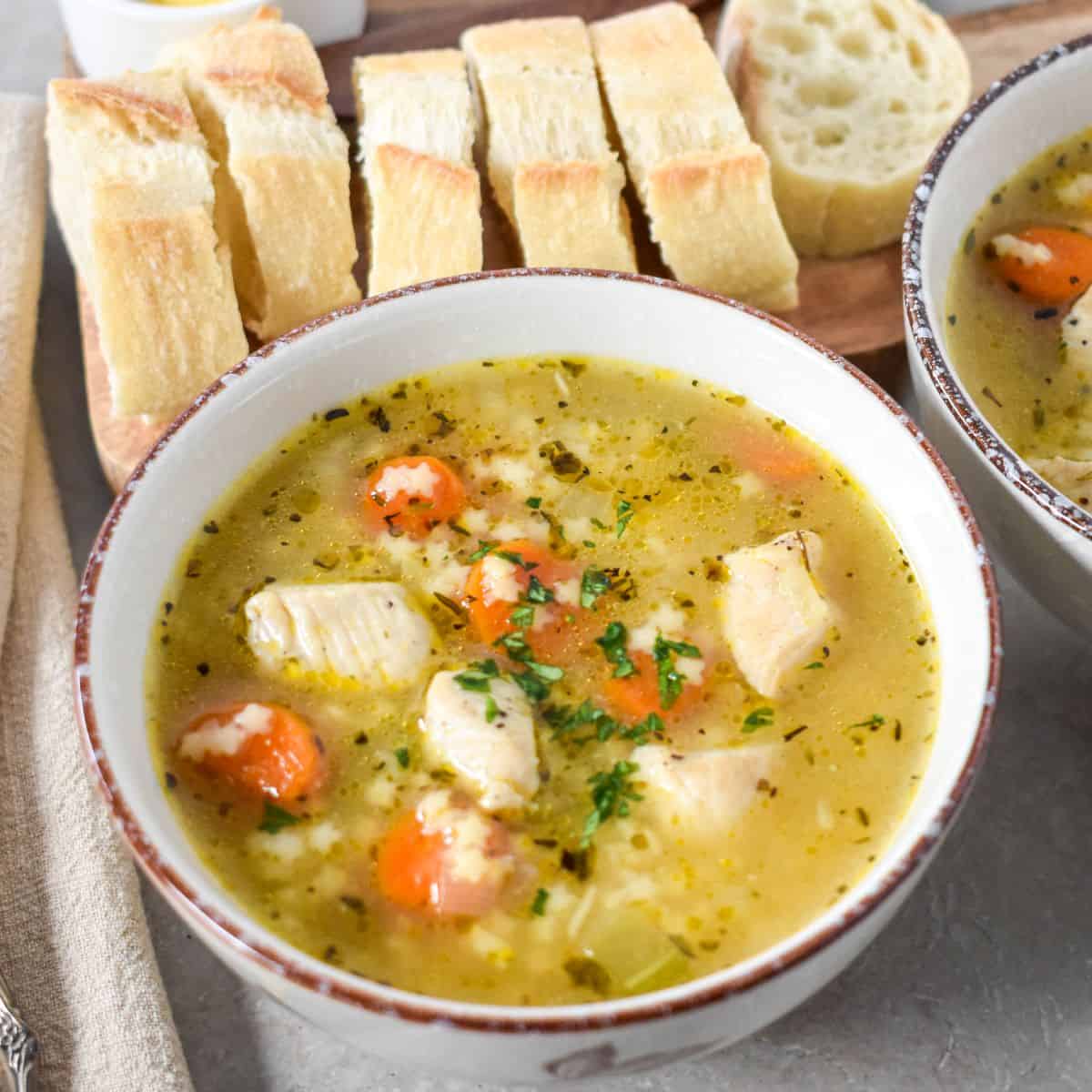 Close-up of the soup in a white bowl with carrots, chicken, and stelline pasta, served with sliced bread on a wooden board.