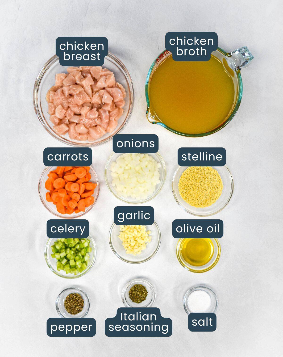 The labeled ingredients for the soup in small bowls, including chicken breast, chicken broth, carrots, celery, onions, garlic, stelline pasta, olive oil, Italian seasoning, salt, and pepper.