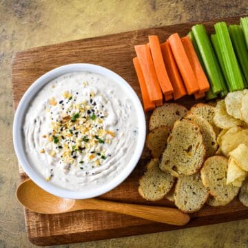 Bowl of everything bagel dip garnished with seasoning and chives, surrounded by vegetables, chips, and crostini on a wooden board.