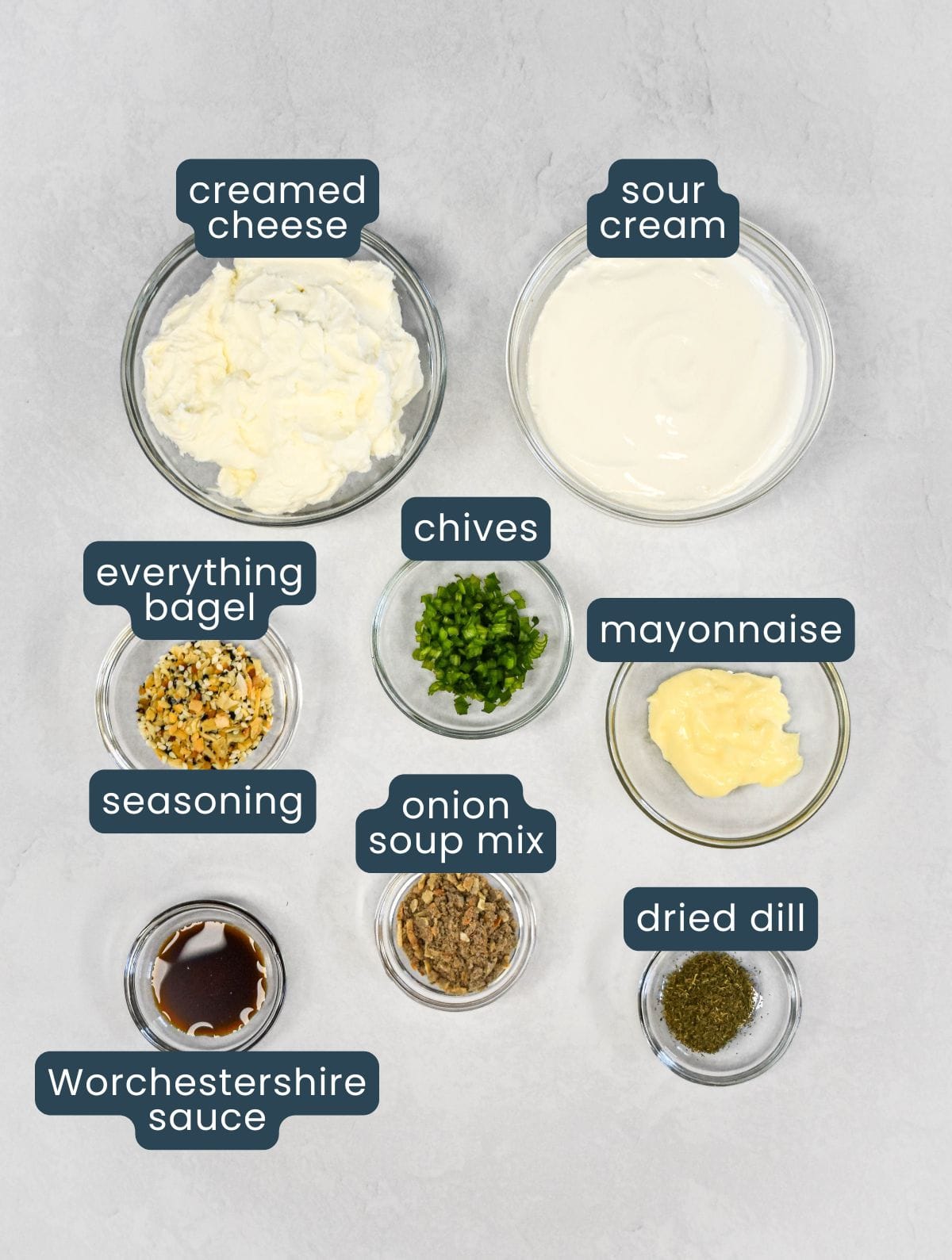 Overhead view of ingredients in small glass bowls, including cream cheese, sour cream, mayonnaise, chives, dried dill, onion soup mix, seasoning, and Worcestershire sauce.