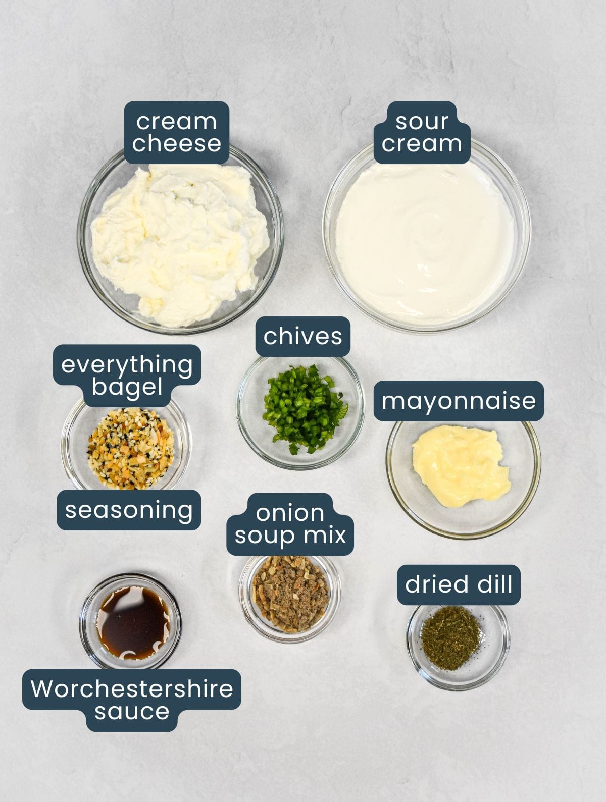 Overhead view of ingredients in small glass bowls, including cream cheese, sour cream, mayonnaise, chives, dried dill, onion soup mix, seasoning, and Worcestershire sauce.