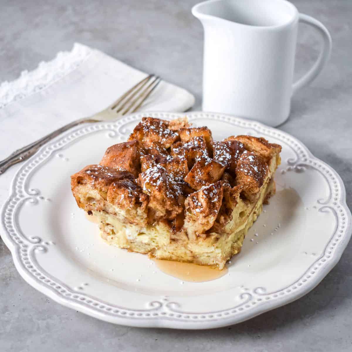 .Slice of baked casserole on a white plate with syrup and a small pitcher in the background.