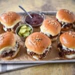 Shredded beef sliders arranged on a baking sheet with pickles and barbecue sauce.
