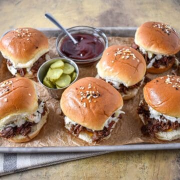 Shredded beef sliders arranged on a baking sheet with pickles and barbecue sauce.