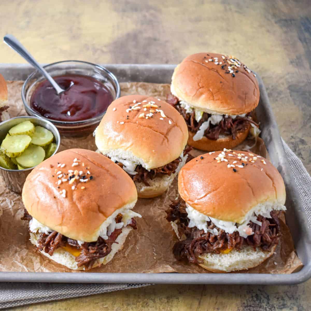 A tray of small beef sandwiches topped with coleslaw, served with pickles and a bowl of barbecue sauce.