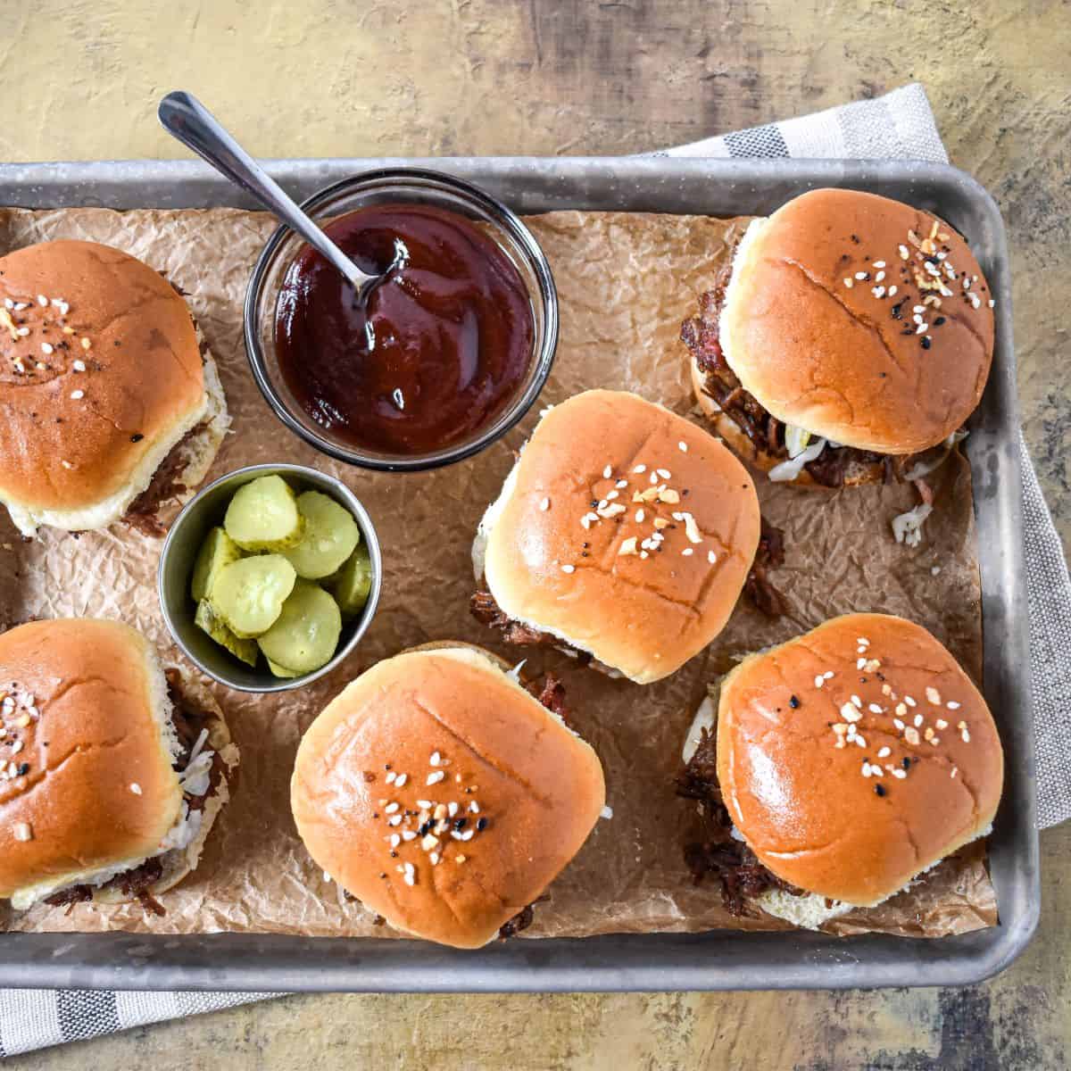 Overhead view of several assembled sliders on a parchment-lined baking sheet with small bowls of pickles and barbecue sauce.