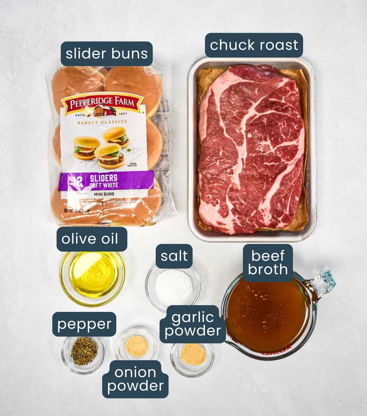 Ingredients for the sliders arranged on a white surface with each item labeled, including the chuck roast, slider buns, seasonings, olive oil, and beef broth.