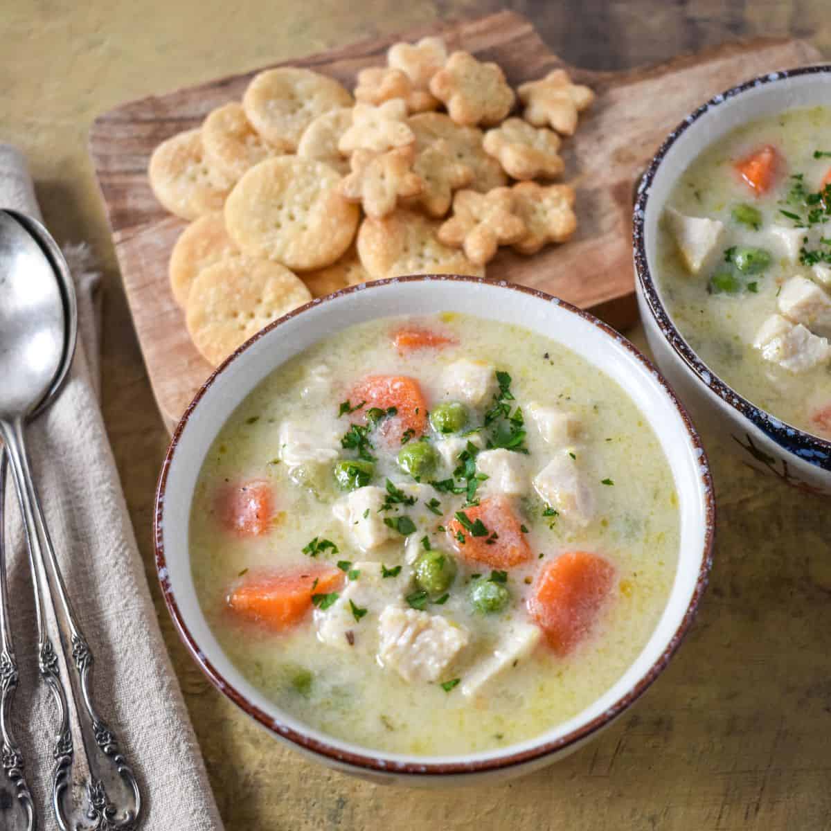 A bowl of creamy vegetable and turkey soup with carrots, peas, and herbs, served with crackers on a wooden board.