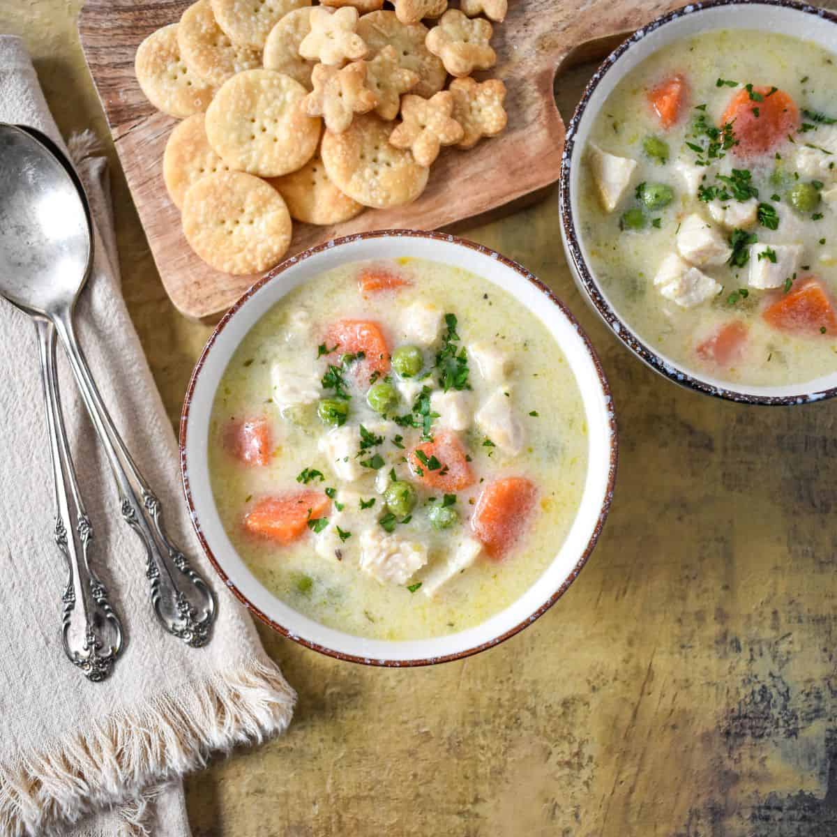 Two bowls of creamy turkey and vegetable soup on a rustic surface with crackers and spoons on the side.