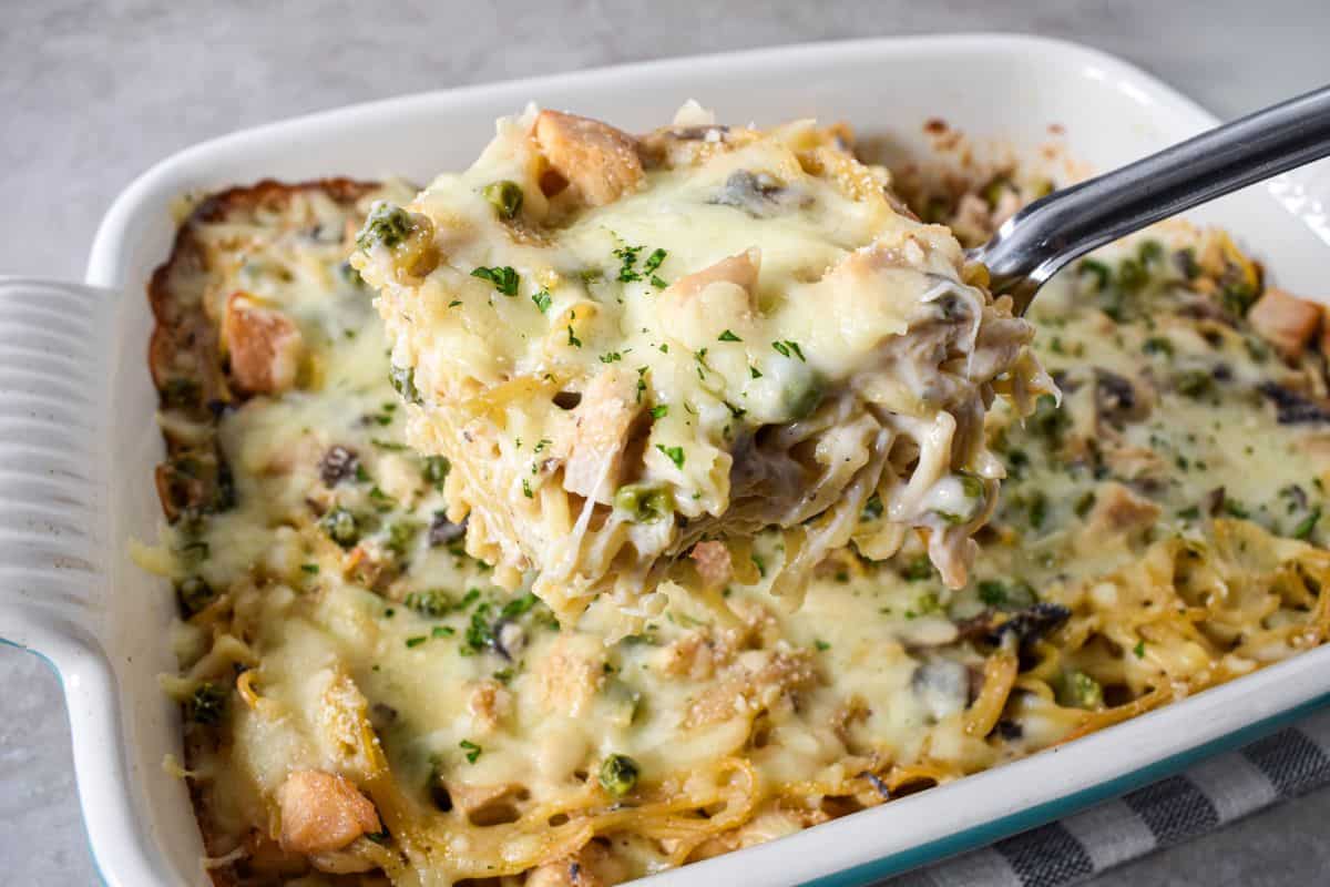 Spoon lifting a cheesy portion of baked casserole from the dish, showing noodles, turkey, peas, and mushrooms.
