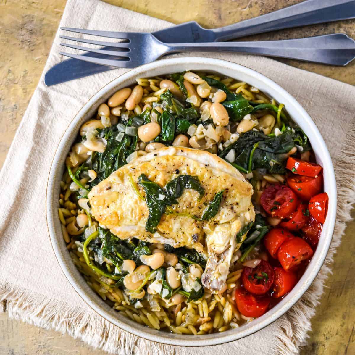 Airline chicken with cannellini beans served over orzo with wilted greens and cherry tomatoes in a shallow bowl.