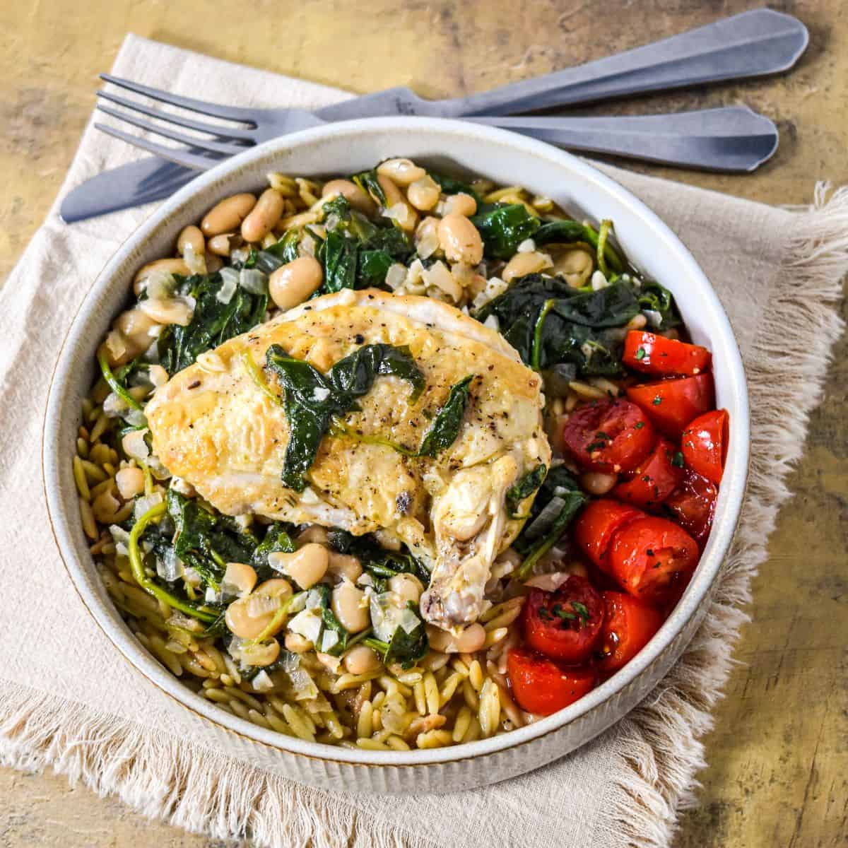 Skillet airline chicken served with cannellini beans, wilted greens, and orzo in a beige, shallow bowl.