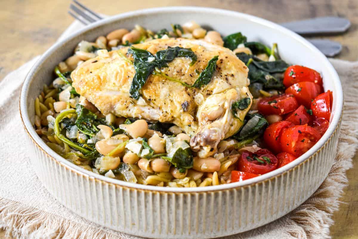 Airline chicken served over orzo with cannellini beans, wilted greens, and cherry tomatoes in a beige, shallow bowl.