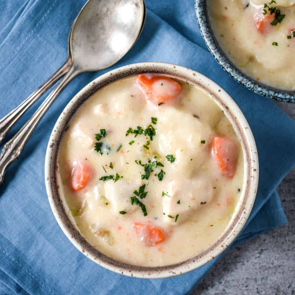 Chicken and dumplings soup served in a rustic ceramic bowl on a blue linen napkin, garnished with parsley, with a second bowl and spoons nearby.