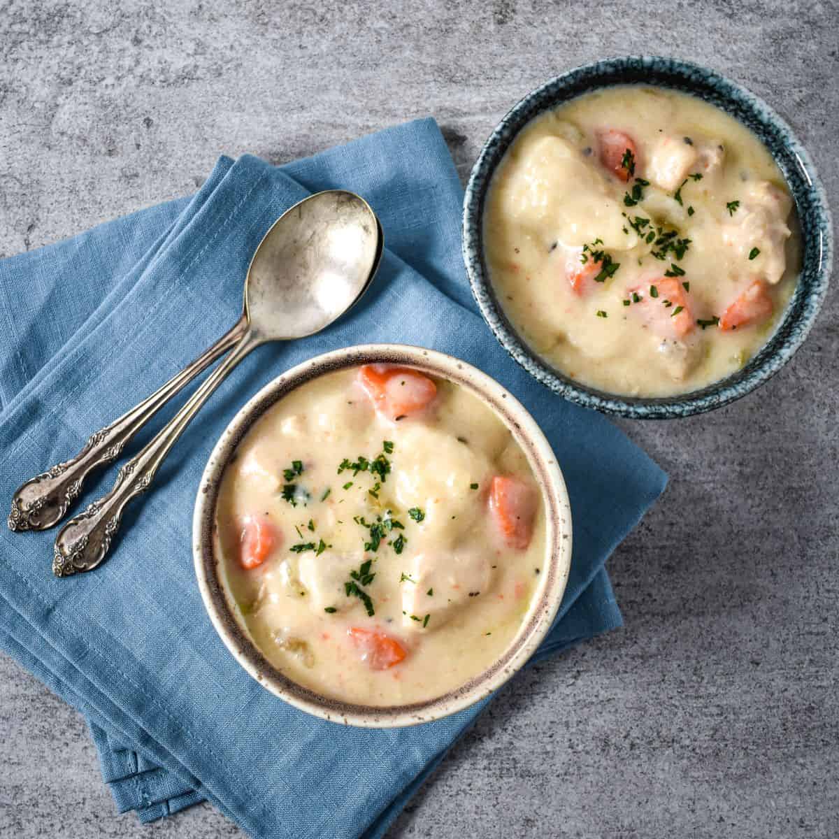 Two bowls of creamy chicken and dumplings soup garnished with parsley, served on a blue napkin with spoons.