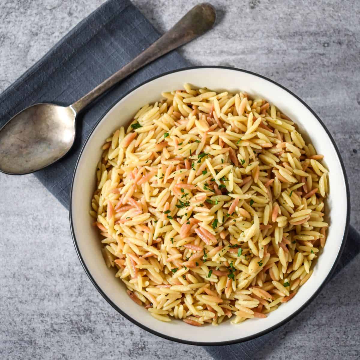 Finished orzo pilaf served in a shallow bowl, set on a gray linen with a serving spoon.