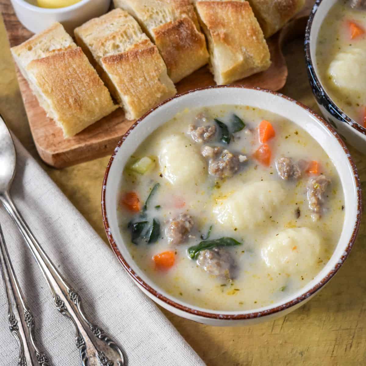 Creamy Italian sausage gnocchi soup with spinach and carrots served in a bowl, with sliced crusty bread on a wooden board.
