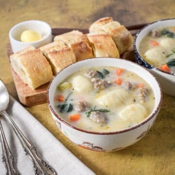 Two bowls of Italian sausage gnocchi soup with spinach and vegetables, served with crusty bread and butter on the side.