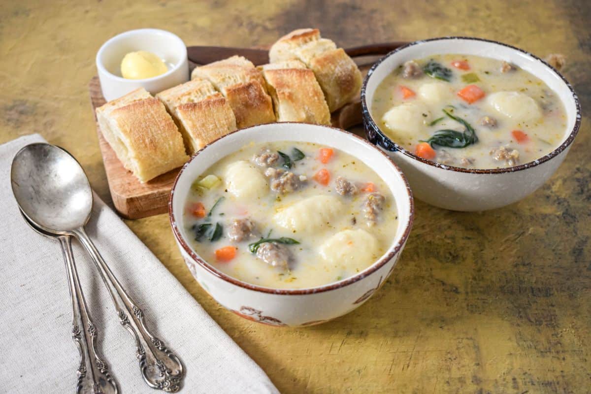 Two bowls of Italian sausage gnocchi soup with spinach and vegetables, served with crusty bread and butter on the side.