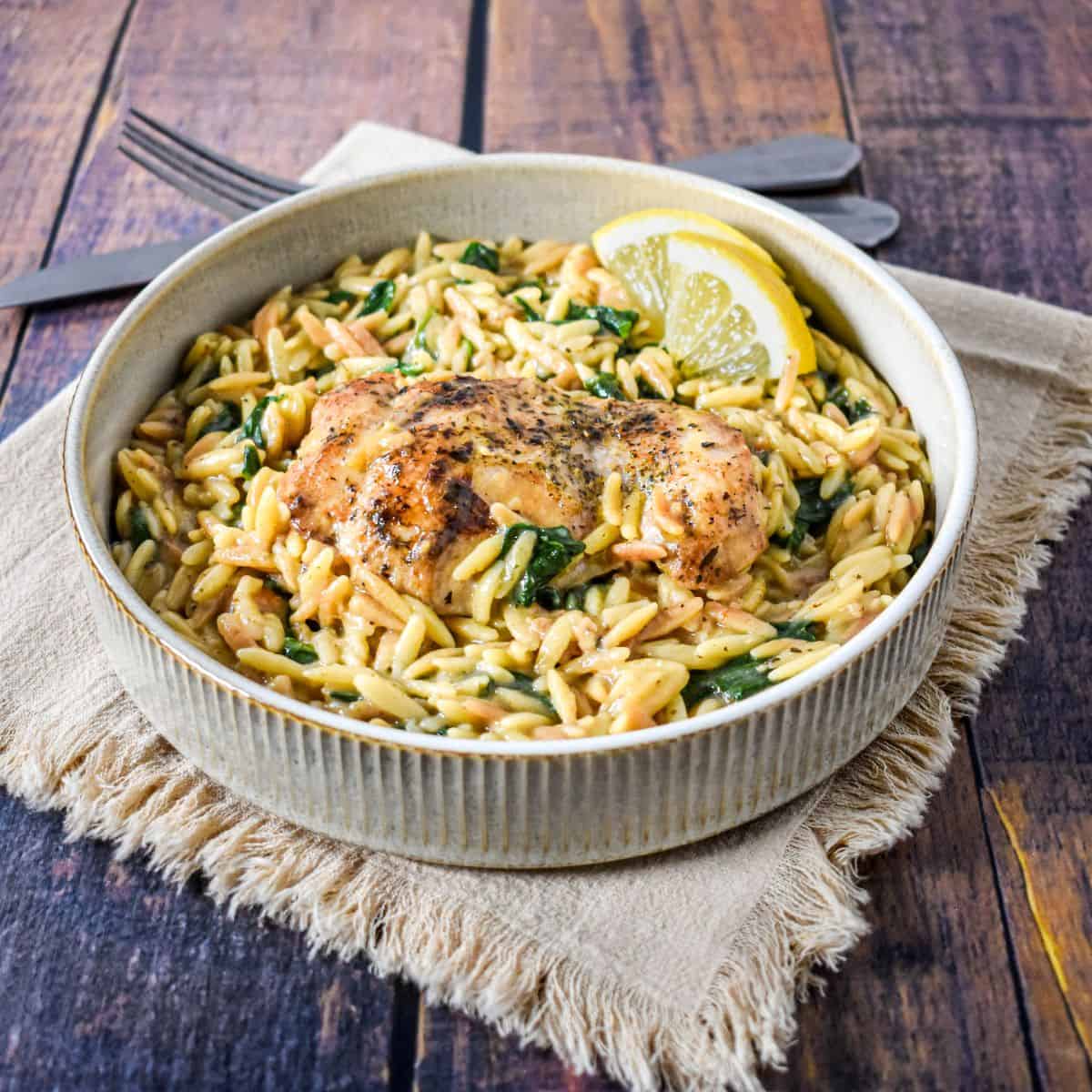 A bowl of chicken, orzo, and spinach garnished with lemon slices, set on a linen napkin.