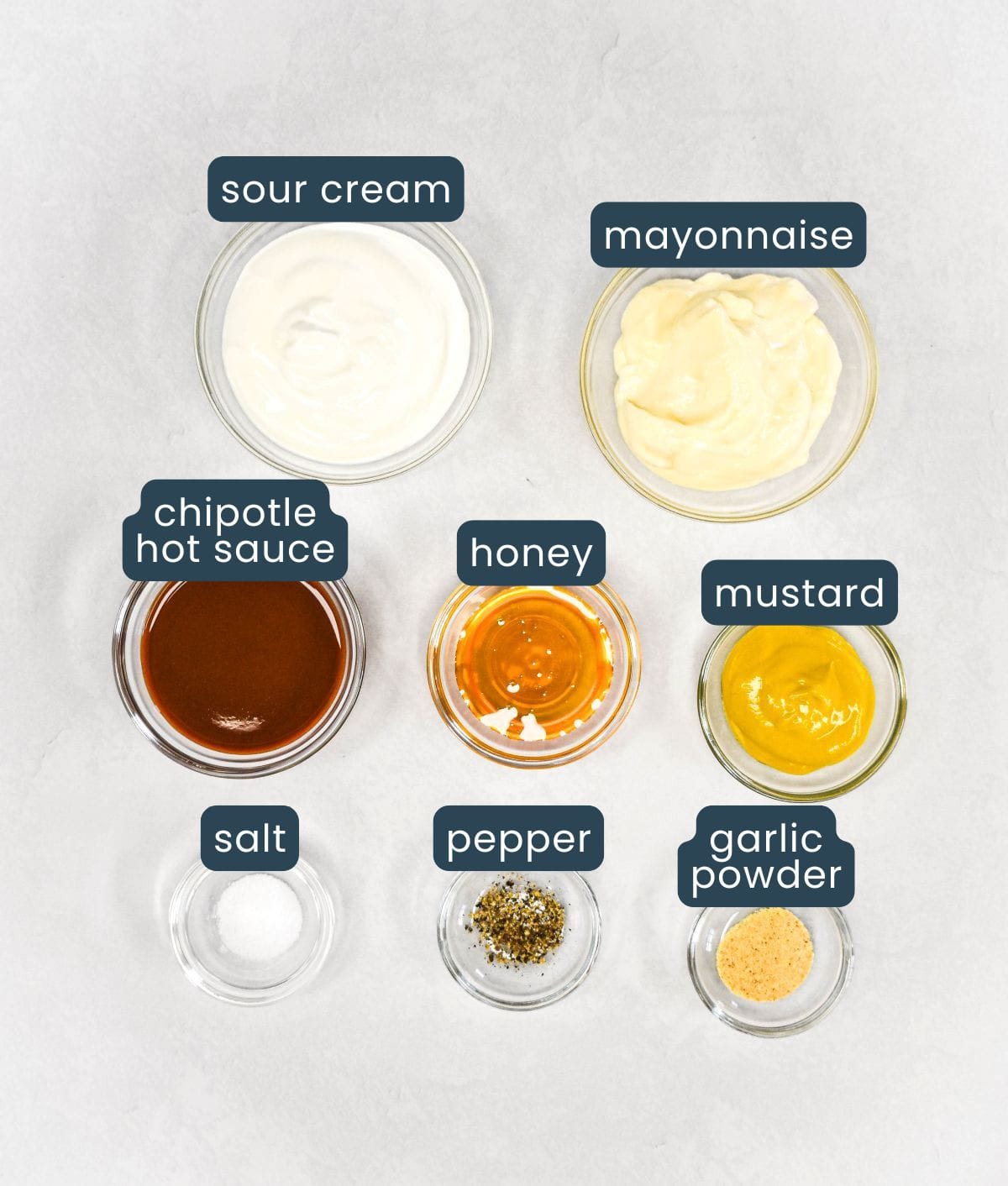 Overhead view of small glass bowls containing sour cream, mayonnaise, chipotle hot sauce, honey, mustard, salt, black pepper, and garlic powder, each labeled for easy identification.
