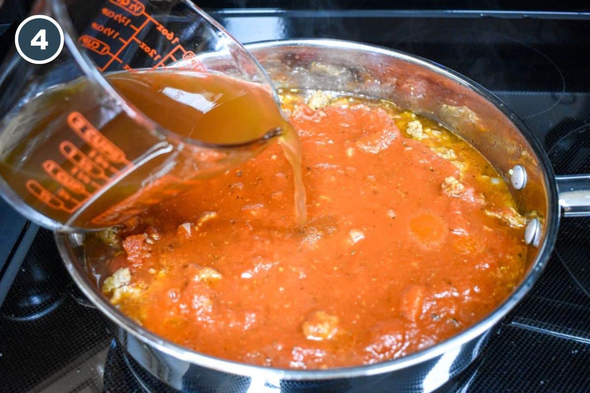 Beef broth being poured from a glass measuring cup into pasta sauce and diced tomatoes in a skillet with sausage.