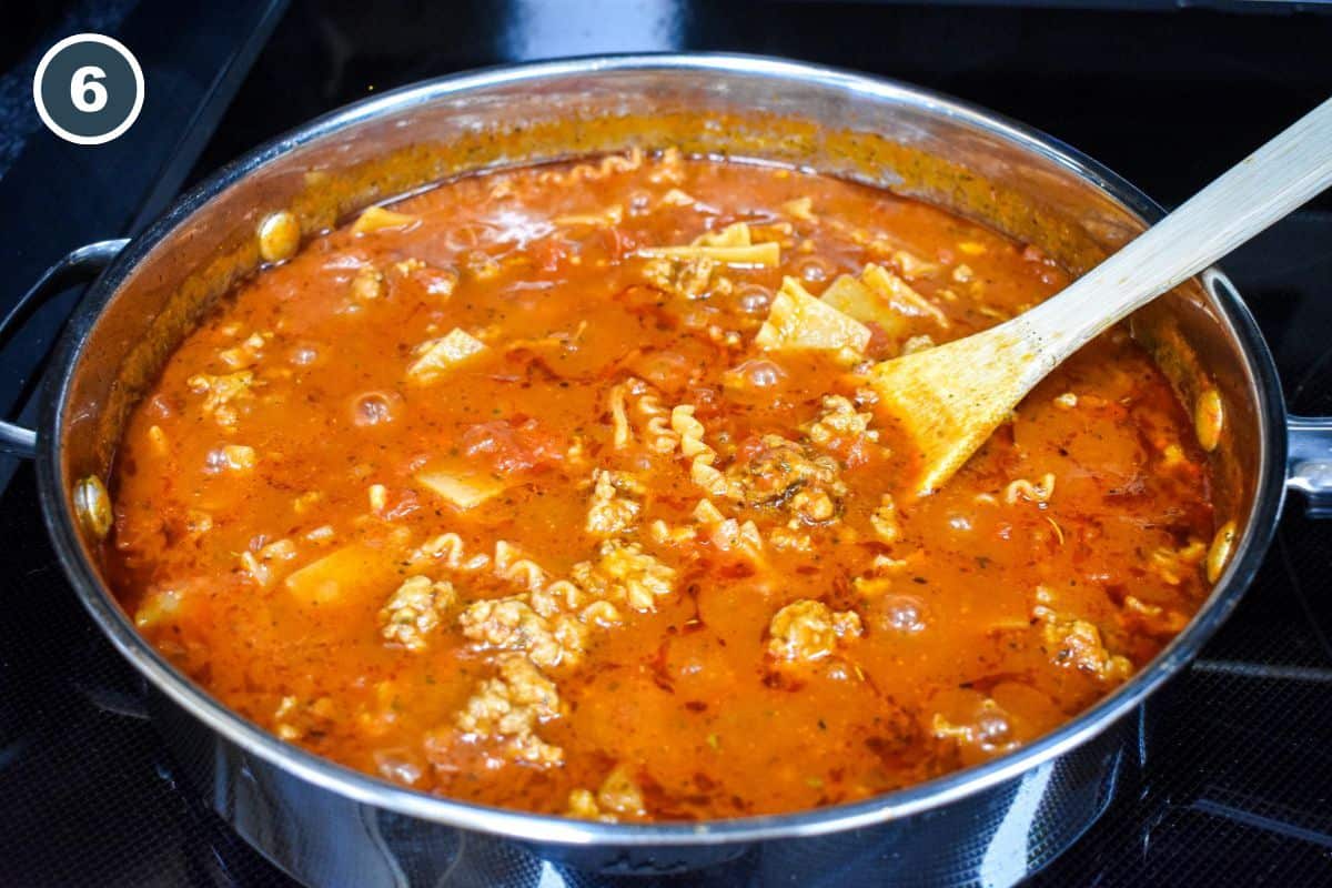 Broken lasagna noodles and sausage simmering in tomato sauce in a skillet, with a wooden spoon resting in the pan.