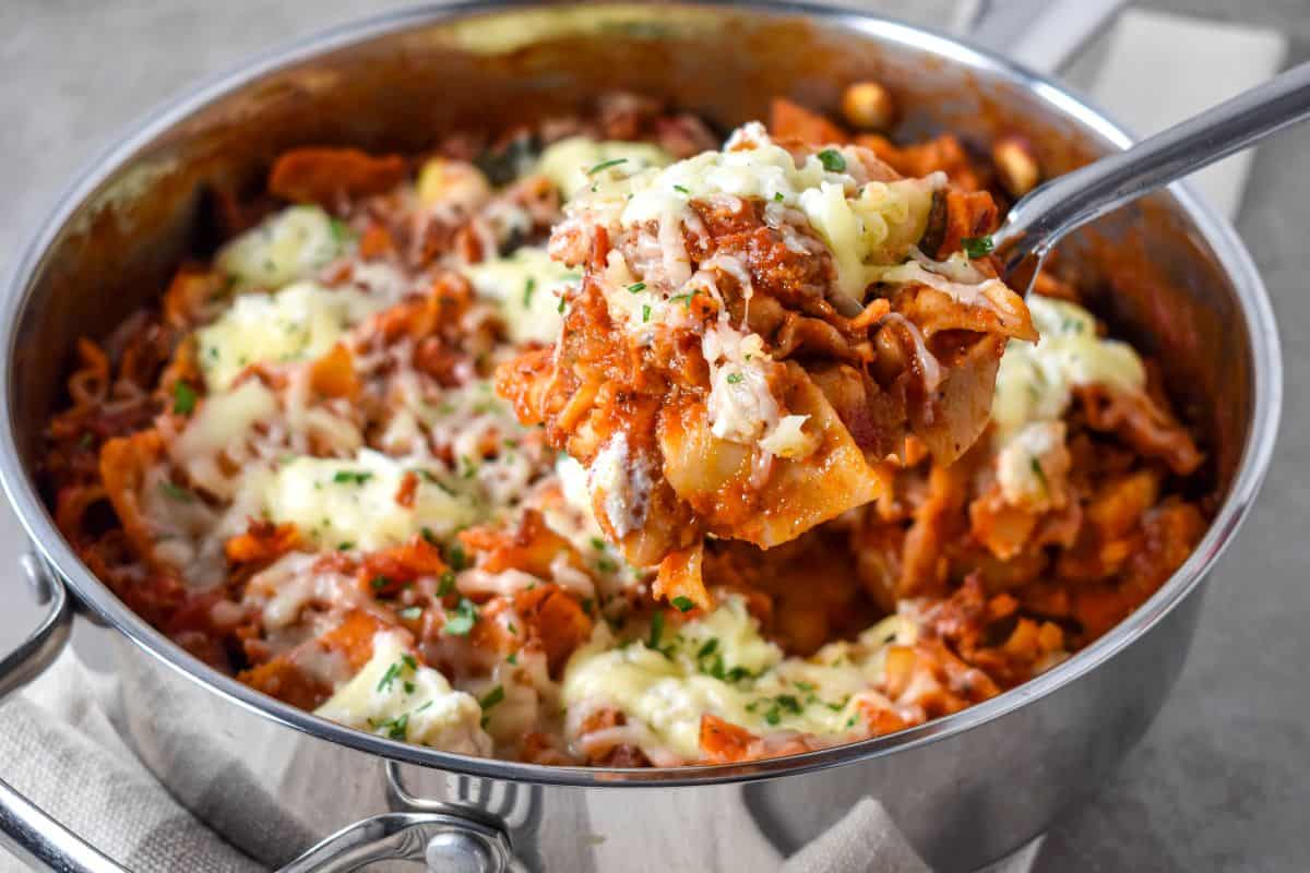 Serving spoon lifting a cheesy bite of skillet lasagna from the pan, with saucy noodles, Italian sausage, and melted mozzarella and ricotta.