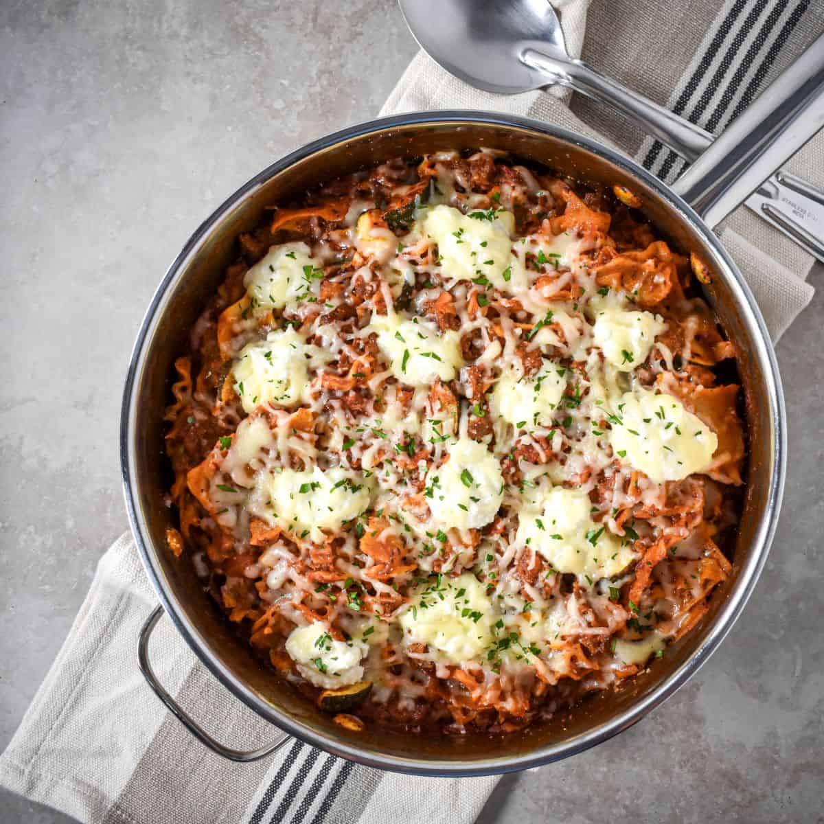 Overhead view of a cheesy sausage and tomato pasta skillet topped with melted mozzarella and dollops of ricotta, garnished with herbs.