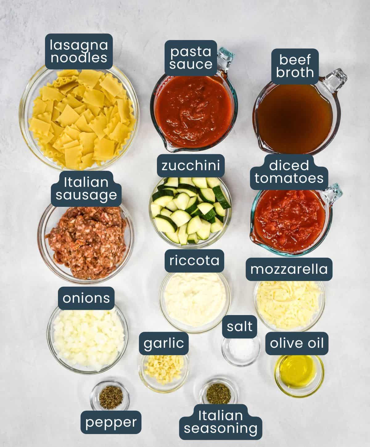 Labeled ingredients for skillet lasagna, prepped and arranged in glass bowls and measuring cups displayed on a white table.