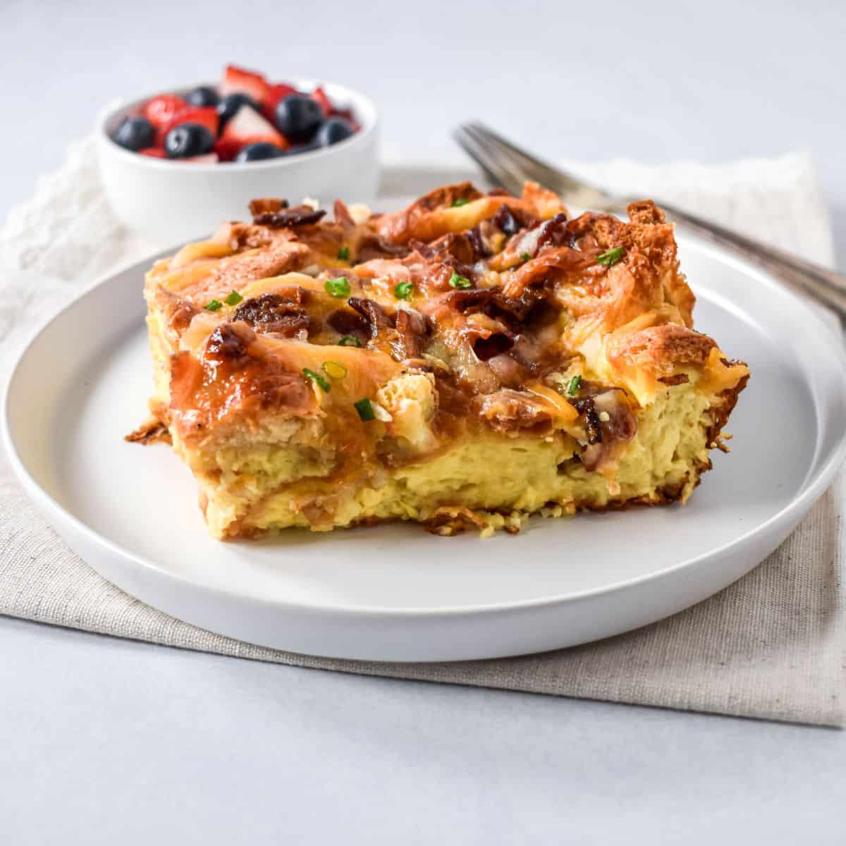 Slice of bacon croissant breakfast casserole on a white plate with fruit in the background.