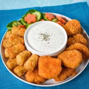 Homemade ranch dressing served in a bowl with chicken nuggets, tater tots, and fresh vegetables on a white plate set on a blue linen.