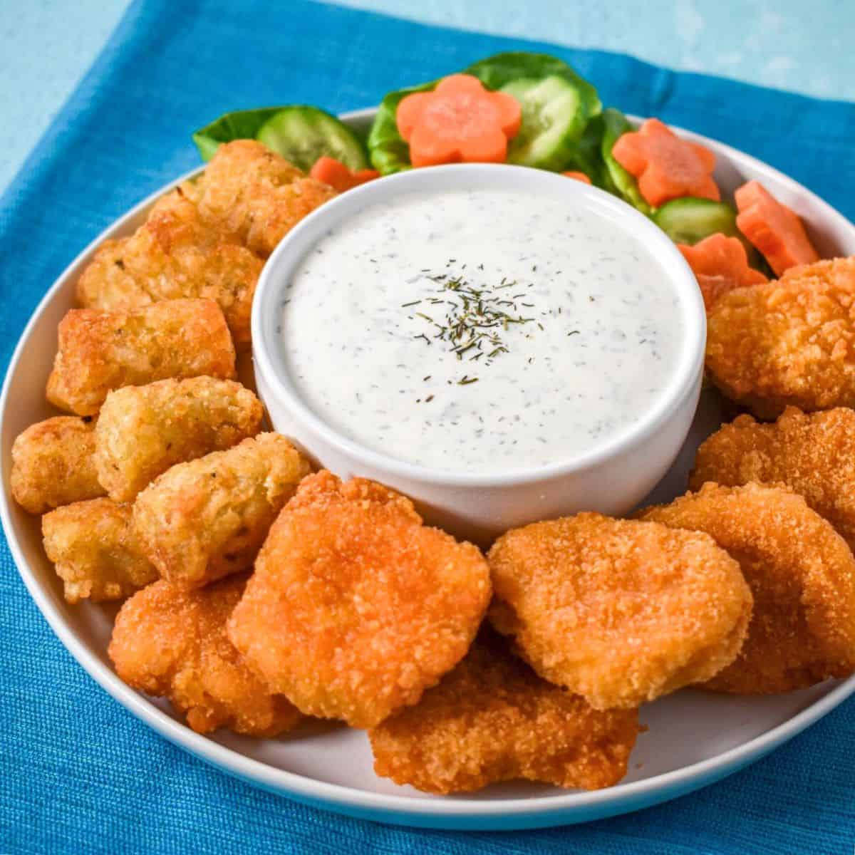 Homemade ranch dressing served in a bowl with chicken nuggets, tater tots, and fresh vegetables on a white plate set on a blue linen.