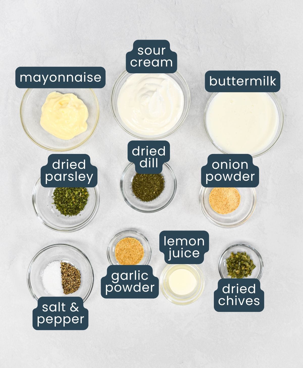 The ingredients for the dressing, including sour cream, buttermilk, mayonnaise, dried herbs, spices, and lemon juice, arranged in small bowls on a white table and labeled.