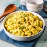 A white bowl filled with creamy deviled egg potato salad, garnished with chopped green onions and paprika, served with a wooden spoon and stacked white bowls in the background.
