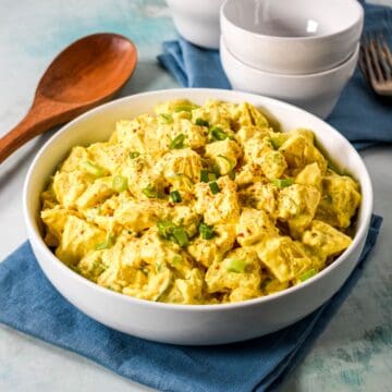 A white bowl filled with creamy deviled egg potato salad, garnished with chopped green onions and paprika, served with a wooden spoon and stacked white bowls in the background.