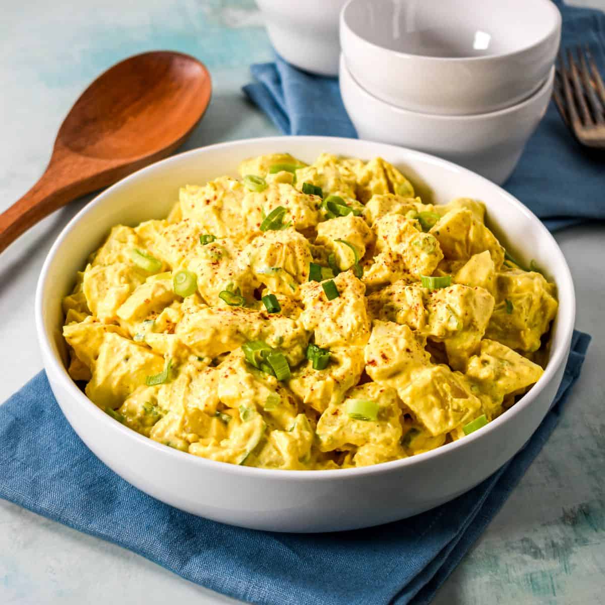 A white bowl filled with creamy deviled egg potato salad, garnished with chopped green onions and paprika, served with a wooden spoon and stacked white bowls in the background.