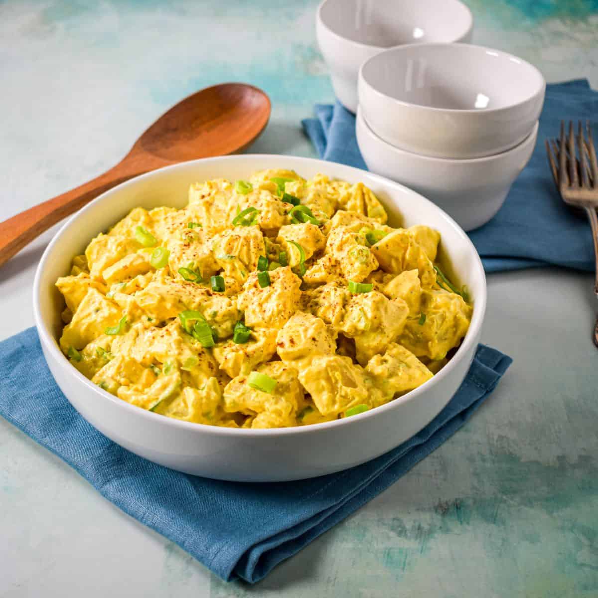 A white bowl filled with creamy deviled egg potato salad, garnished with chopped green onions and paprika, served with a wooden spoon and stacked white bowls in the background.