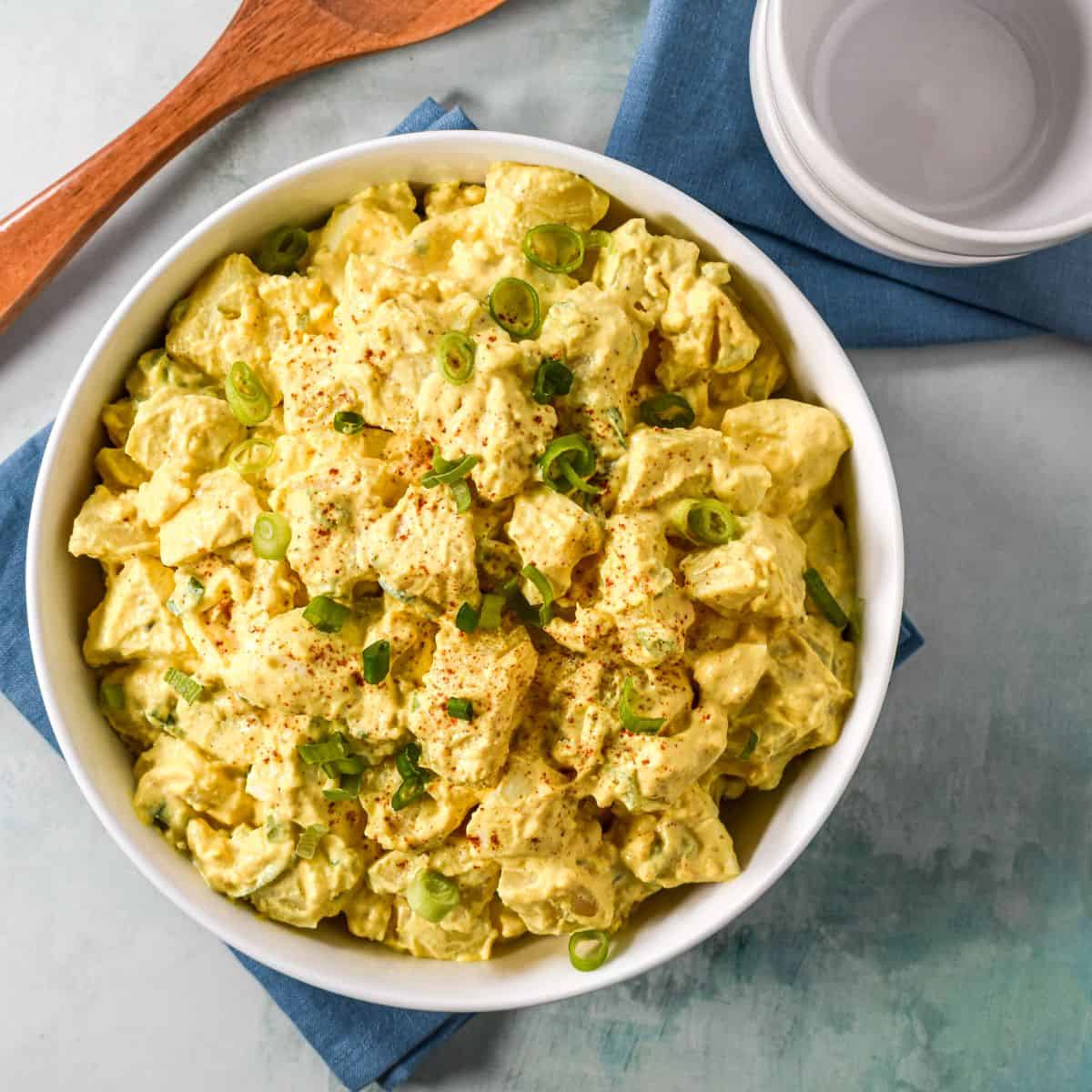 Overhead view of deviled egg potato salad in a white bowl on a blue napkin, styled with a wooden spoon and white bowls.