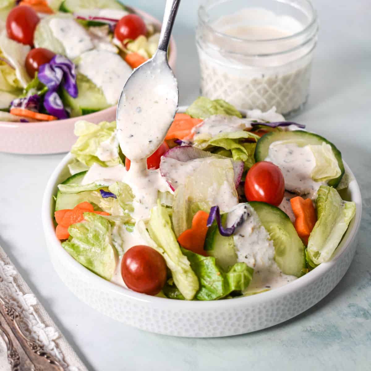 Spoon drizzling parmesan peppercorn dressing over a green salad with tomatoes, cucumbers, and carrots.