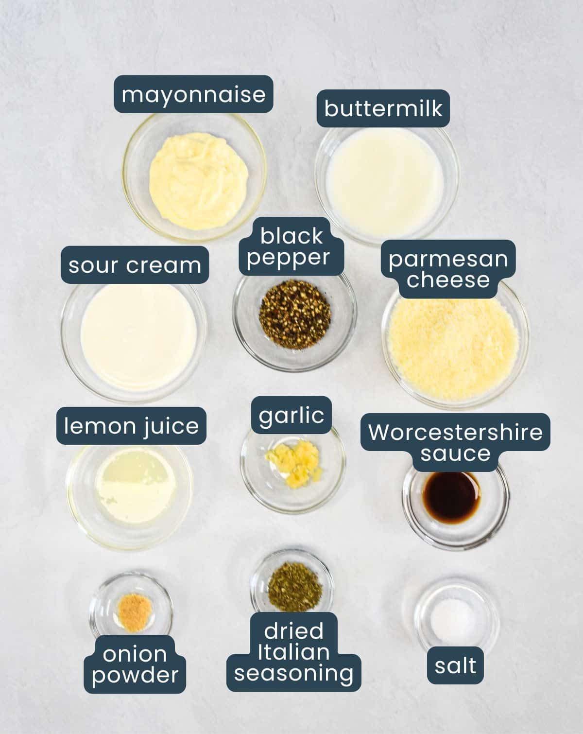 Ingredients for parmesan peppercorn dressing arranged in small glass bowls set on a white table with each one labeled.