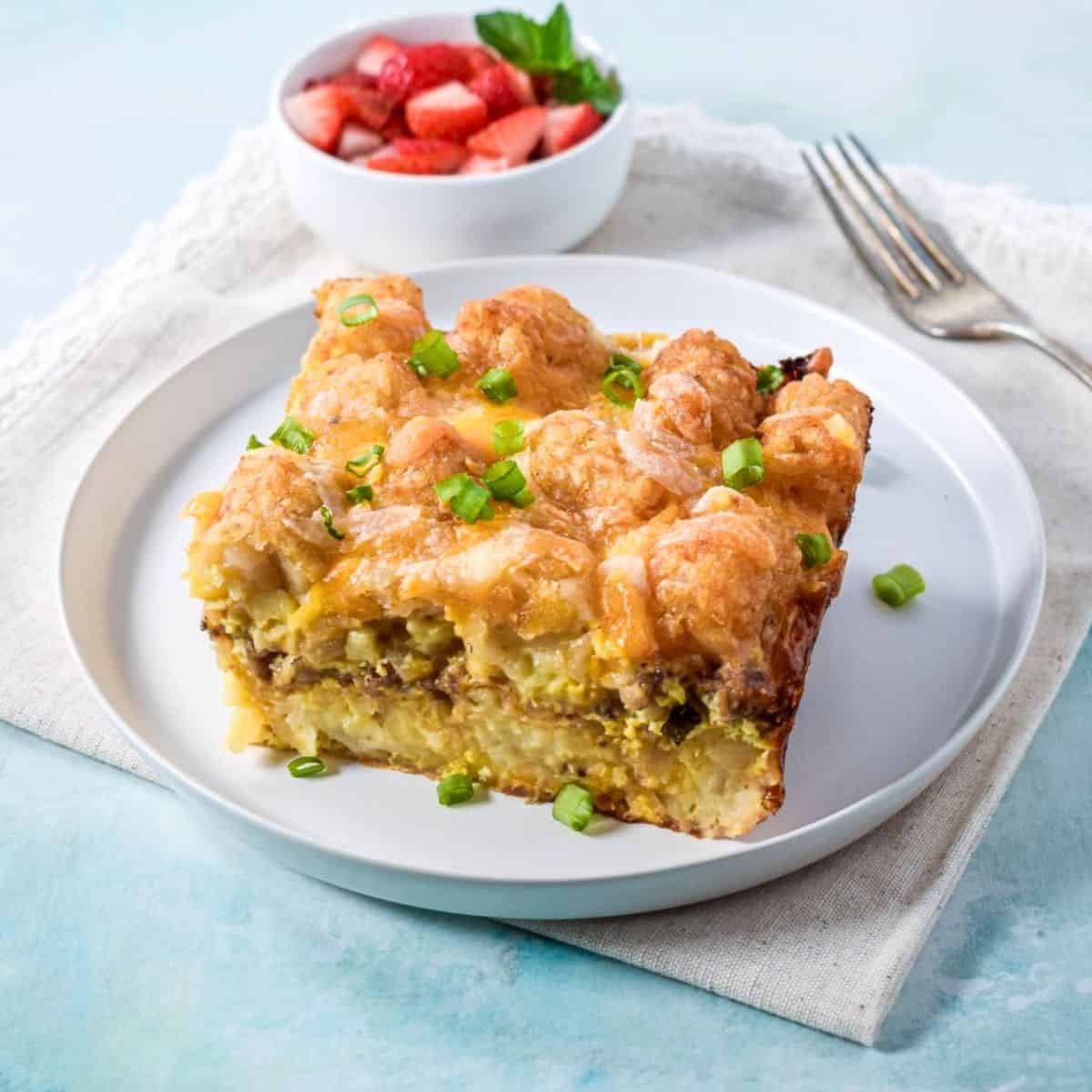 Slice of tater tot breakfast casserole served on a white plate with a bowl of strawberries in the background.
