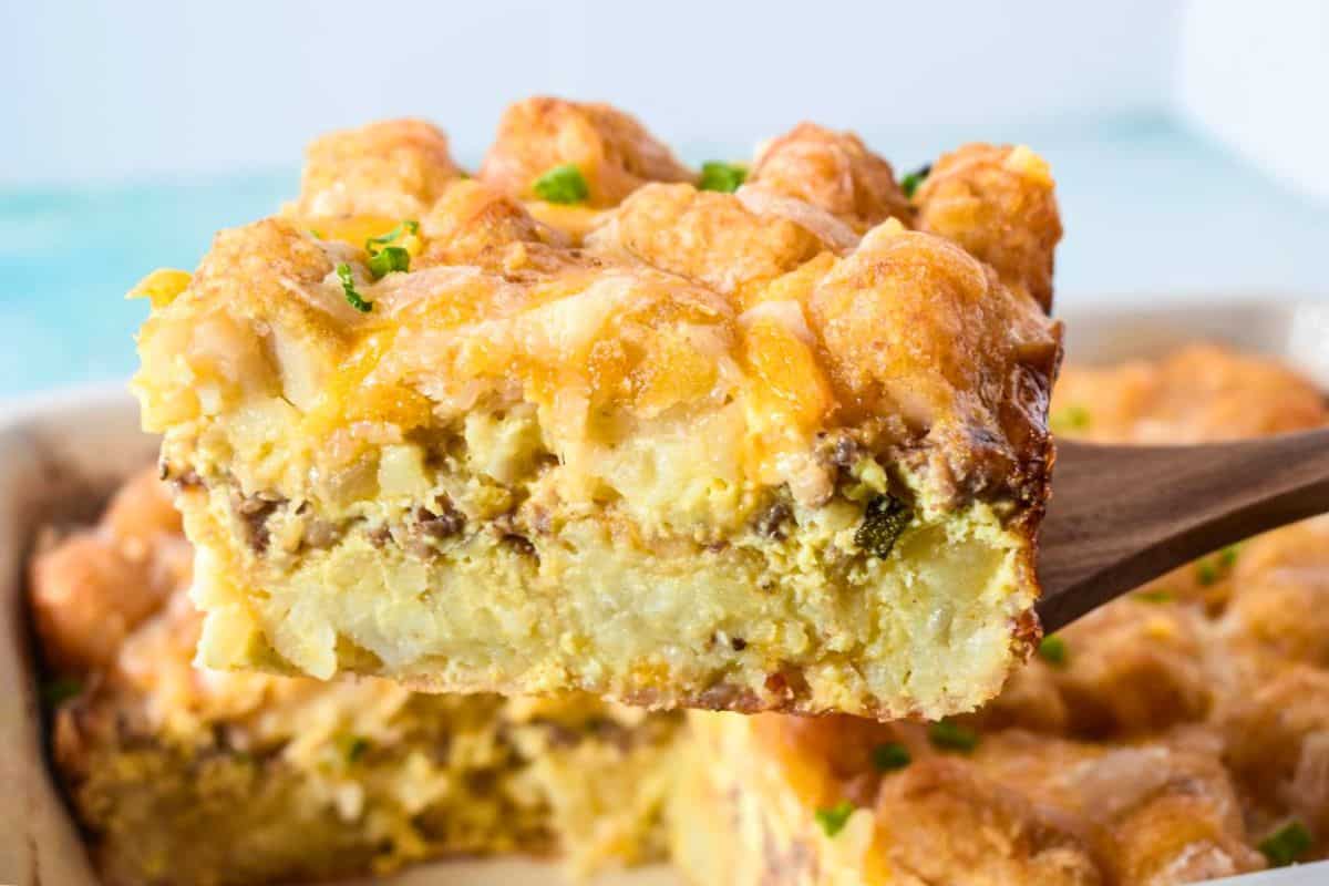 Slice of tater tot breakfast casserole on a wooden spatula showing the layers of eggs, sausage, cheese, and tater tots.