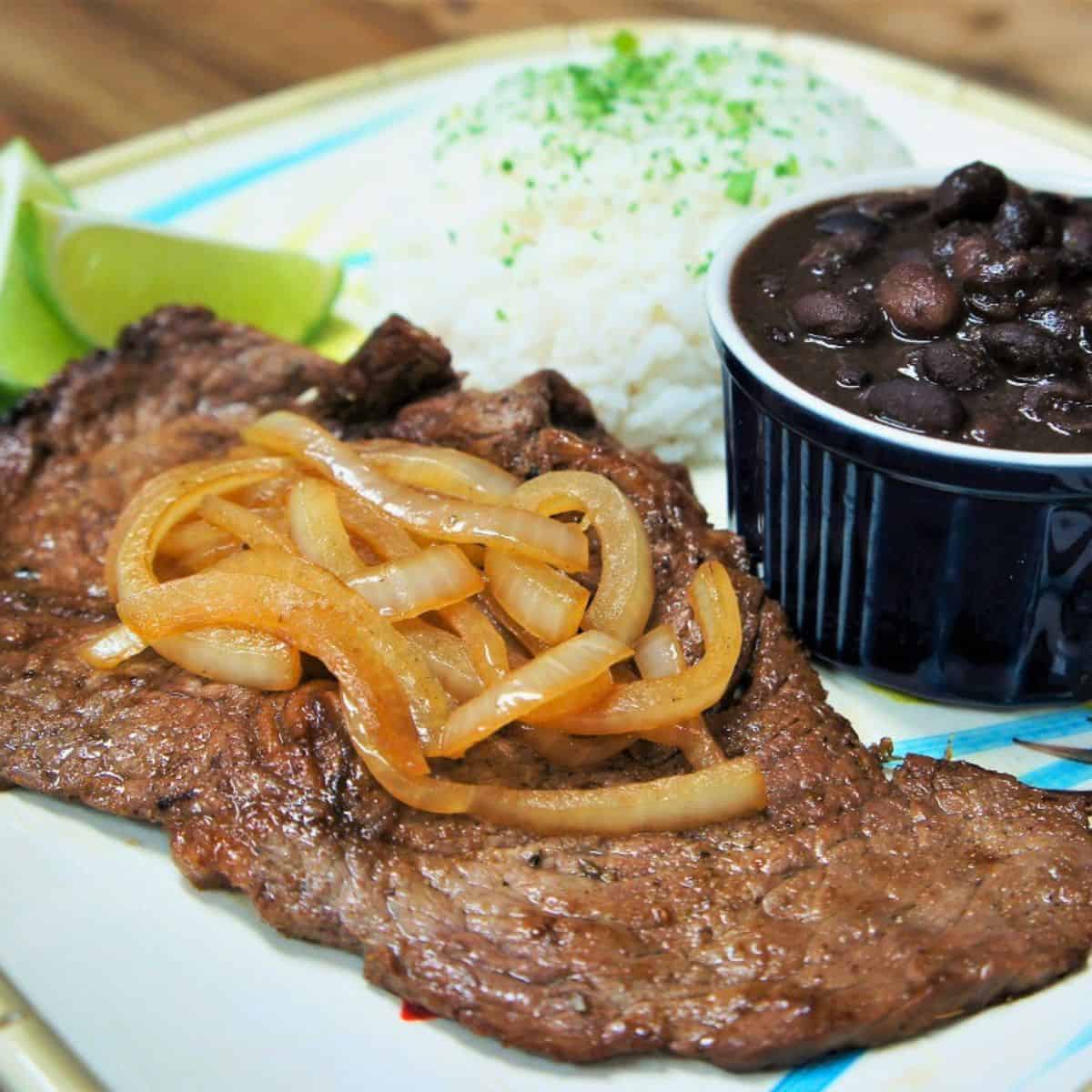 Bistec de Palomilla, thin sirloin steak served with cooked onions, white rice and black beans.
