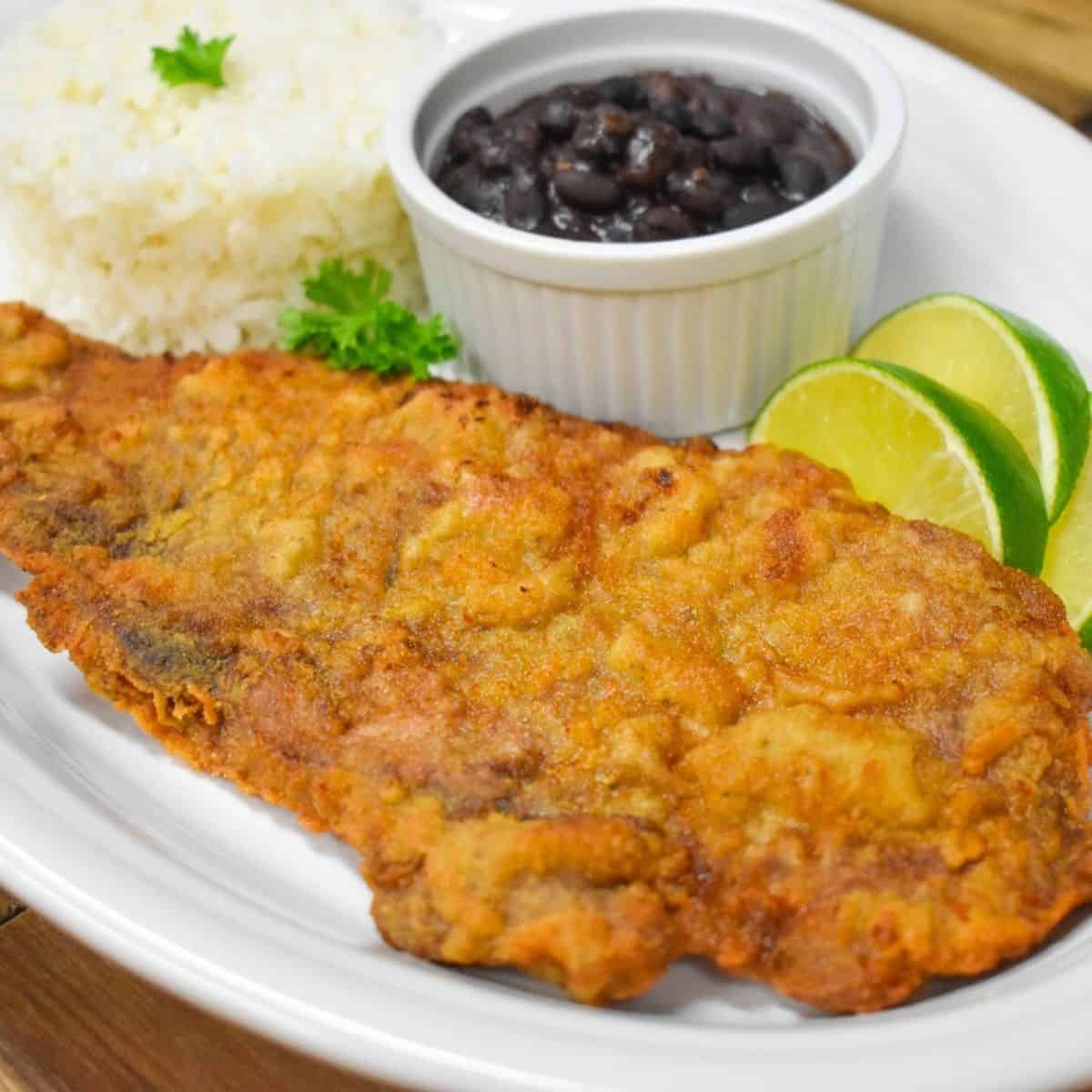 A bistec empanizado, served on a large white platter with white rice and blacks on the side and garnished with lime wedges.
