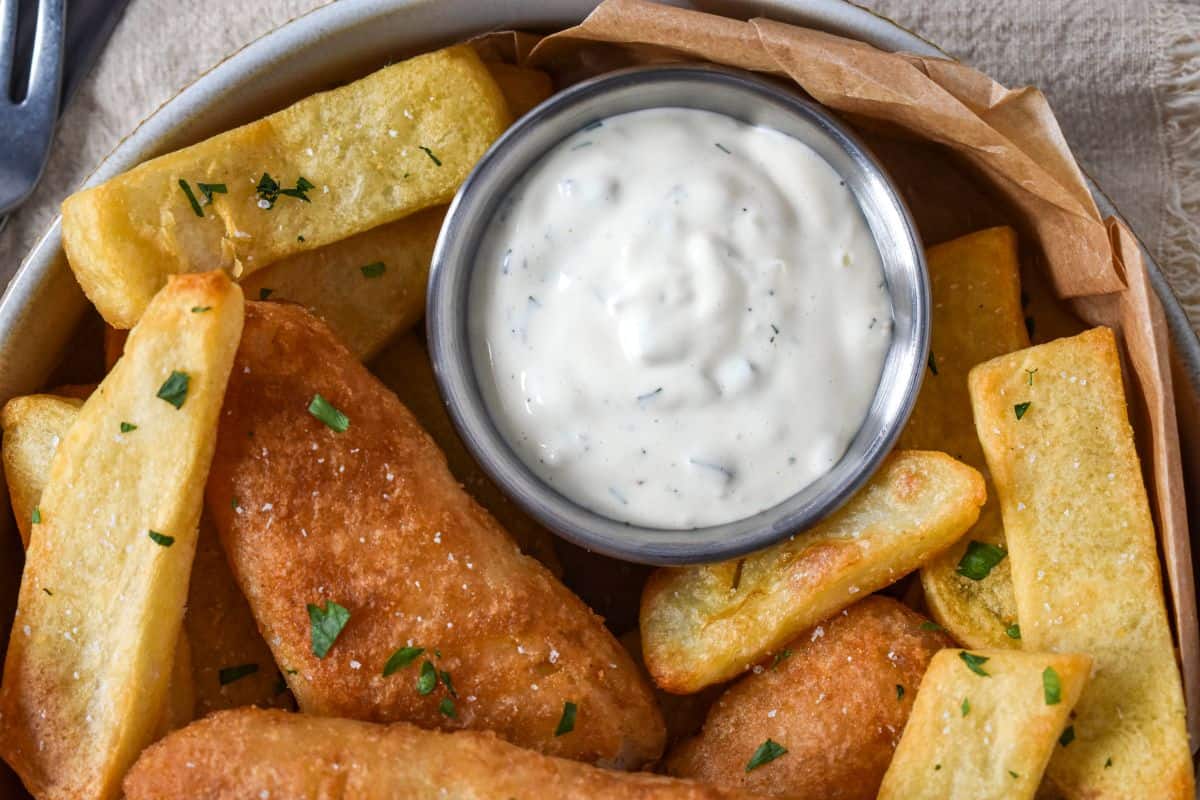 Close-up of tartar sauce in a small metal cup surrounded by battered fish and French fries.
