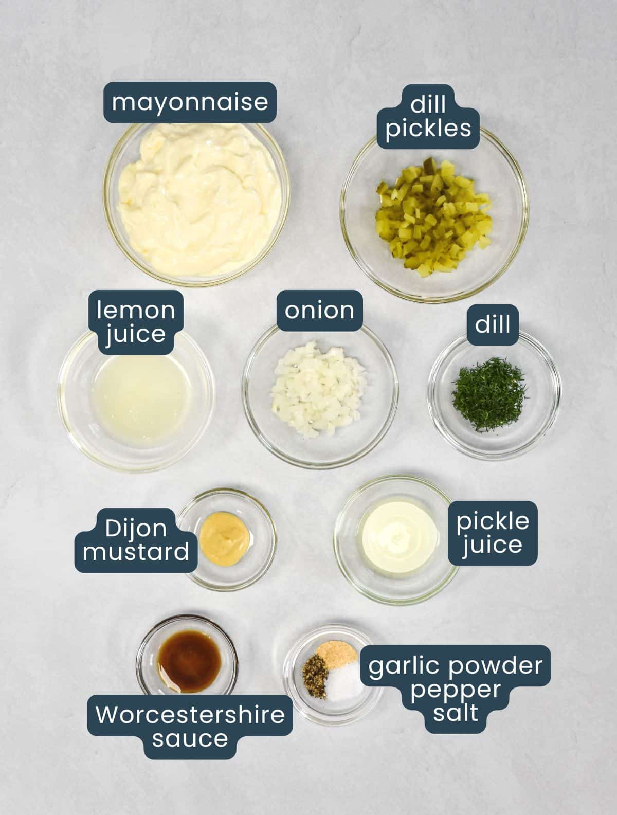Ingredients for tartar sauce arranged in small bowls on a white background.